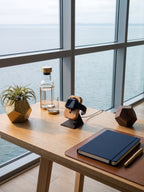 KI-generiertes Bild: Ein heller Holztisch an einem Fenster mit Blick auf das Meer, darauf eine Apple Watch auf einem Holz-Ladeständer, ein Wasserkrug, eine Sukkulente im Topf und ein Notizbuch mit Stift.